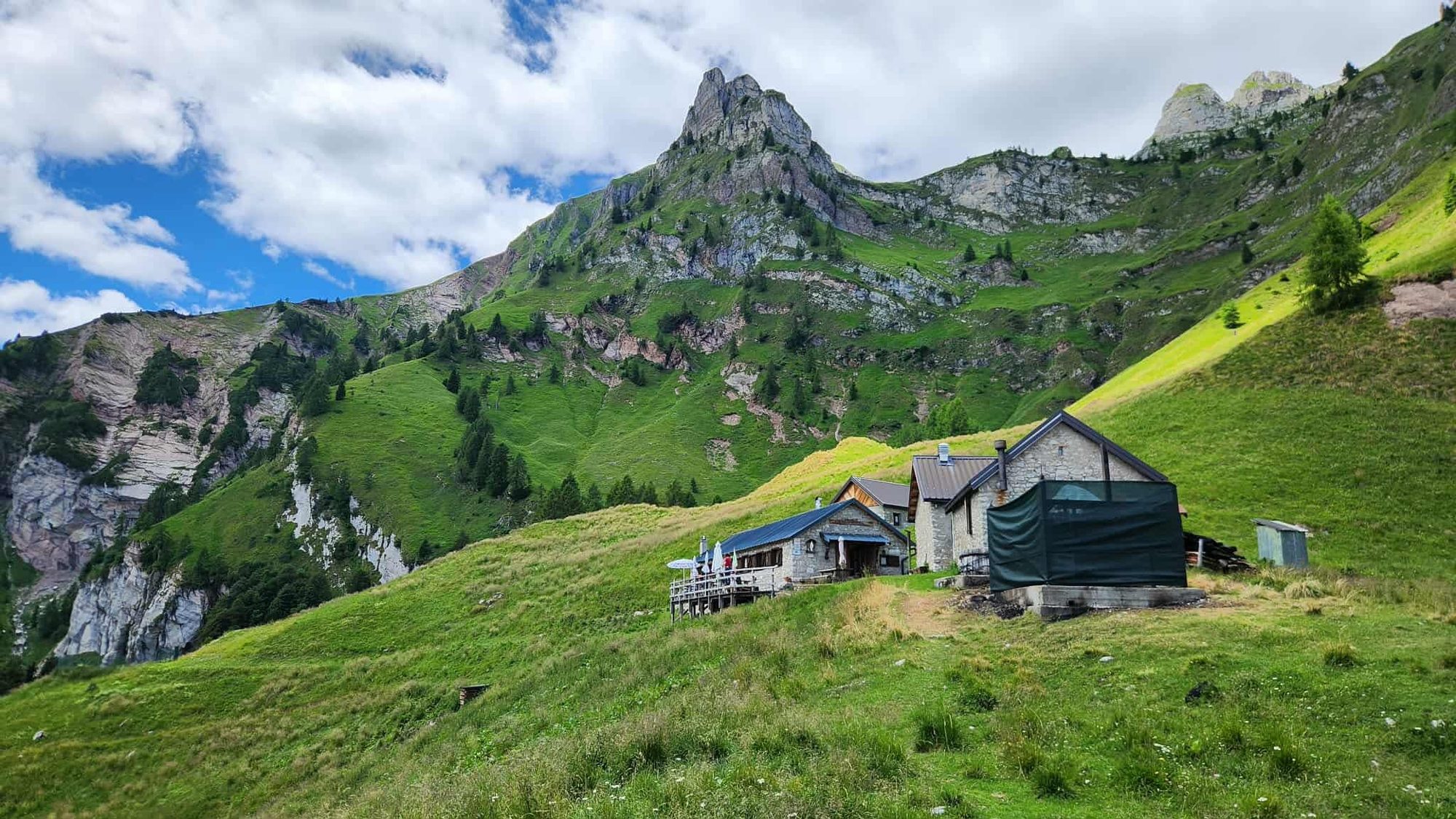 A mountain hut below green Dolomite slopes and rocky cliffs.