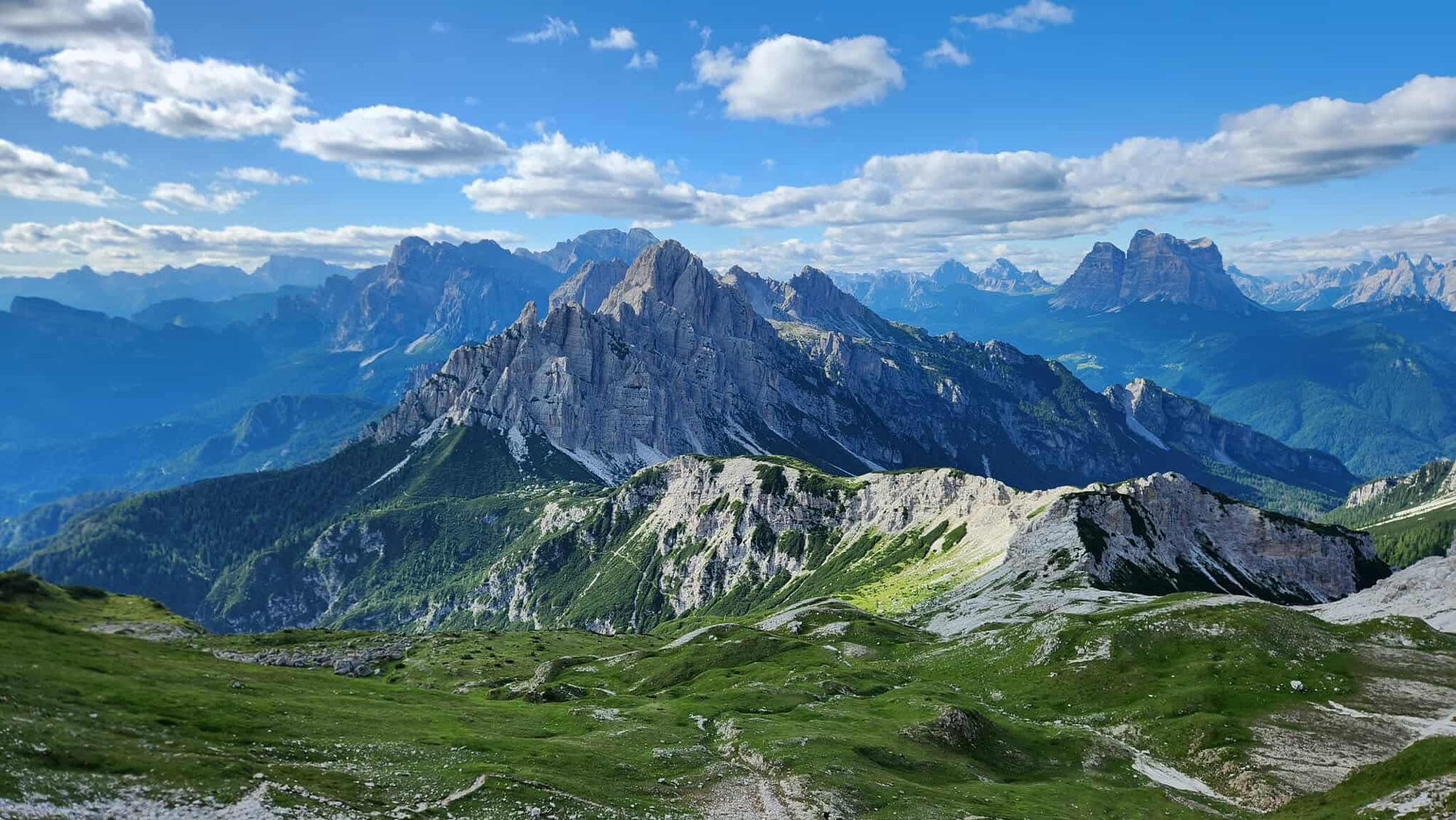 Wide panorama of jagged Dolomite peaks under a blue sky above green alpine terrain.